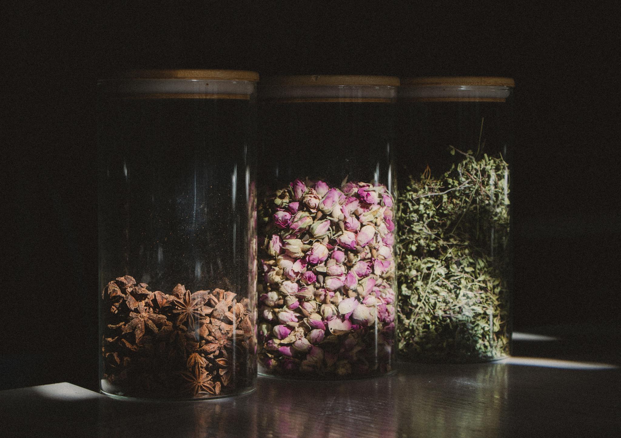Three glass jars containing star anise, dried rosebuds, and herbs beautifully lit.